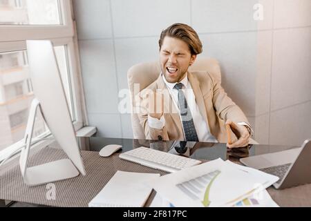 Overjoyed male employee in stylish formal suit is sitting at the desk, feeling euphoric, got a job promotion. Happy businessman is using computer Stock Photo