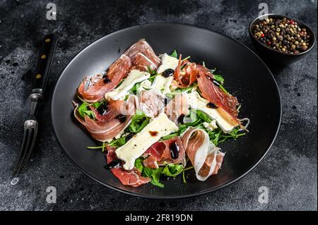 Prosciutto crudo ham salad with brie camembert cheese and arugula on a plate. Black background. Top view Stock Photo