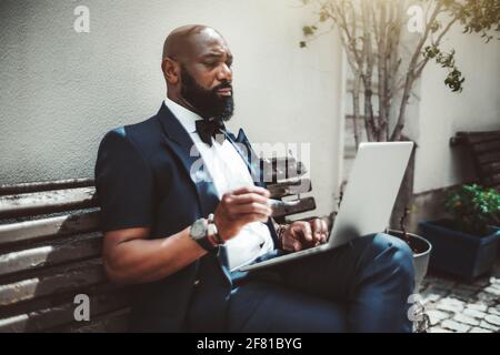 A fancy handsome bald bearded African senior with glasses in a hand is working on a netbook outdoors; portrait of a black man entrepreneur with a well Stock Photo