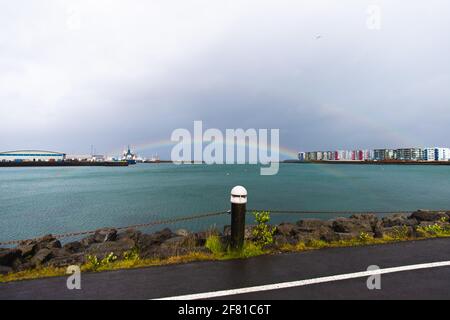 complete rainbow over a lake on a very cloudy day next to a road Stock Photo
