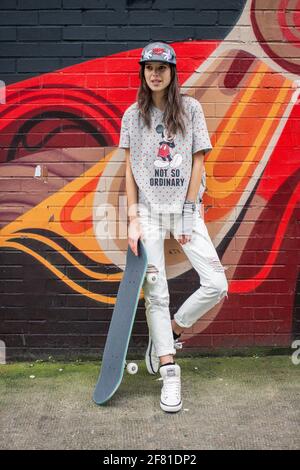 Teenage girl with skateboard standing near by urban wall with colorful graffiti in Shorditch in London , UK . Stock Photo