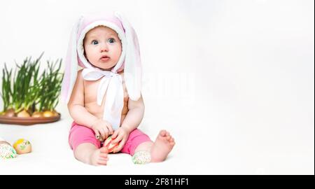 A little girl with the ears of a pachy rabbit sits on a bed near colorful eggs. The funny child looks up in surprise. religion concept Stock Photo