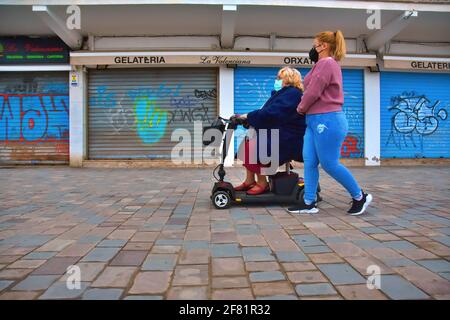 Calafell, Spain. 10th Apr, 2021. A woman in an electric wheel chair and the other walking, move past a closed ice cream parlour during the local confinement in Calafell. The Government of Catalonia once again orders the regional confinement due to the Covid-19 health crisis, reducing the mobility of people resulting to empty streets, beaches, restaurants, cafes and ice cream parlours being closed at 5:00 pm. Credit: SOPA Images Limited/Alamy Live News Stock Photo