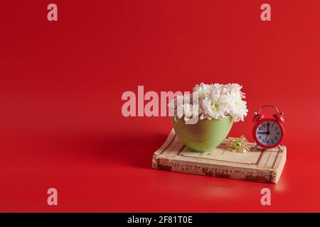 on a closed book there are white flowers in a green cup with an alarm clock  Stock Photo