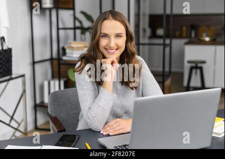 Executive female employee in casual wear sitting at the office desk in front of the laptop, friendly woman looks at the camera and smiles, balanced and tranquility freelancer woman on the workplace Stock Photo