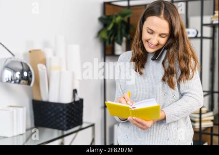 Cheerful woman has phone conversation and writes down into notebook, standing in the office, multitasking female office employee Stock Photo