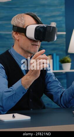 Retired man experiencing virtual reality using vr headset in living room working from home. Old remote employee searching, analysing financial reports while elderly wife watching tv in background Stock Photo