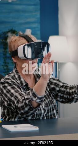 Retired woman experiencing virtual reality using vr headset in living room working from home. Old remote employee searching, analysing financial reports while elderly wife watching tv in background Stock Photo