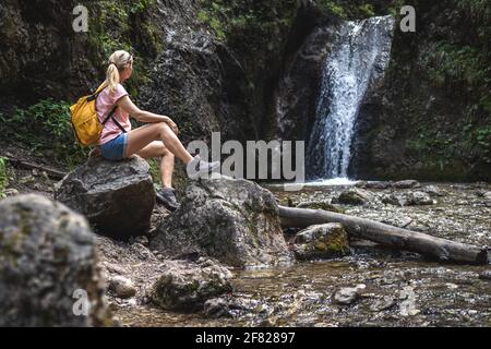 Woman resting during hike in wonderful nature. Hiker sitting on rock near stream with waterfall in forest at natural parkland Mala Fatra, Slovakia Stock Photo