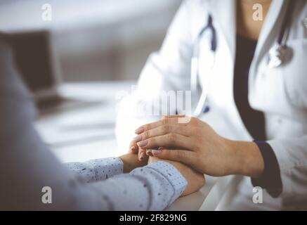 Unknown woman-doctor is holding her patient's hands, discussing current health examination, while they are sitting at the desk in the cabinet in a Stock Photo
