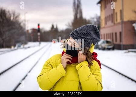 Woman with face mask waiting for train at railroad station in winter. Traveling during coronavirus, covid-19 pandemic. Young woman wearing yellow coat Stock Photo
