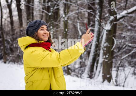 Young woman in winter coat taking selfie by smartphone during walk outdoors. Smiling caucasian woman using mobile phone for photo messaging Stock Photo