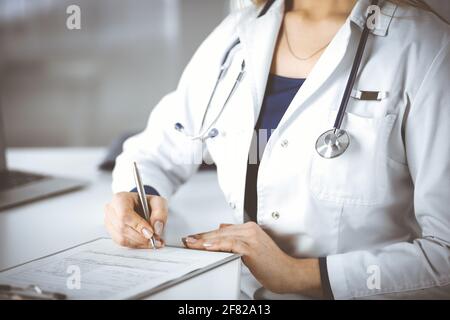 Unknown woman-doctor is prescribing some medication for her patient, using a clipboard, while sitting at the desk in her cabinet. Female physician Stock Photo