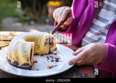 Senior woman cutting slice of bundt cake outdoors. Refreshment in spring garden. Serving Easter cake on table Stock Photo