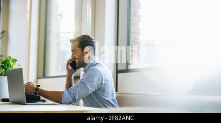 businessman talking with client on mobile phone. Young male sitting at office desk with laptop talking on smart phone. Stock Photo