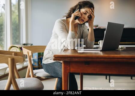 Woman having headache talking on cellphone while working from home. Woman sitting at table with laptop computer looking stressed out while talking ove Stock Photo