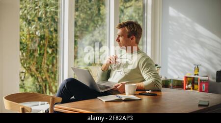 Thoughtful man with laptop at home. Man sitting by table with laptop and looking away thinking. Stock Photo