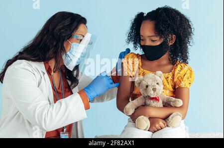 Woman in medical uniform giving injection to a girl wearing face mask in clinic. Doctor giving covid-19 vaccine to a girl during pandemic. Stock Photo