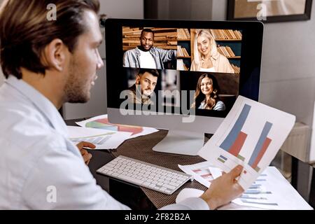 Serious male employee is having an online meeting with coworkers, discussing financial graphs. Stylish businessman is using computer, distantly Stock Photo