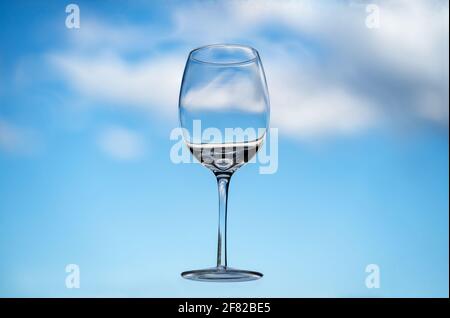 Floating empty wine glass in front of a blue sky with fluffy clouds Stock Photo