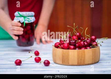 Cherry in wooden bowl and woman holding jar of preserved cherries fruit. Homemade compote in female hands and fresh harvested cherries on table. Stock Photo