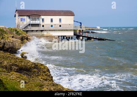 Rough choppy seas crashing on rocks at high tide by new RNLI lifeboat station boathouse in Moelfre, Isle of Anglesey, north Wales, UK, Britain Stock Photo