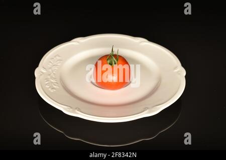 One ripe, bright red, organic, tasty cocktail tomato, close-up, on a white ceramic plate, on a black background. Stock Photo