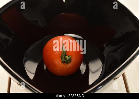 One ripe, bright red, organic, delicious cocktail tomatoe in a black ceramic plate, close-up, on a table made of white painted natural wood Stock Photo