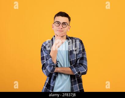Pensive young man in glasses touching his chin, thinking over something on orange studio background Stock Photo