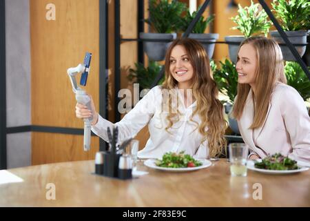 Side view of happy young attractive blogger women eating tasty salads in cafe and communicating with friends online on phone. Concept of free time with delicious food and with good mood.  Stock Photo