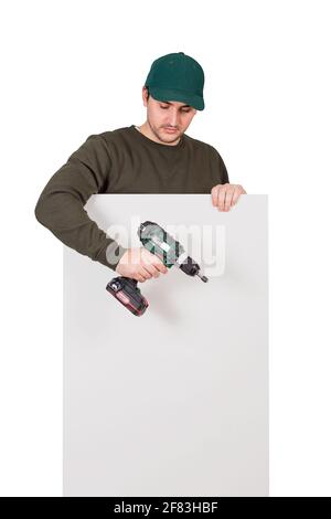 Young man with a screwdriver or electric drill in his hand, stands behind a white panel. Renovation worker before installing interior finish plate or Stock Photo