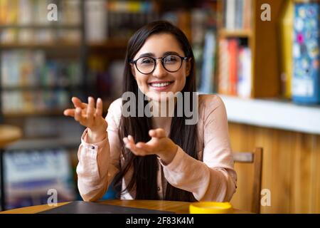 Remote tutoring concept. Young teacher talking and gesturing at camera, sitting in modern cafe, working distantly Stock Photo