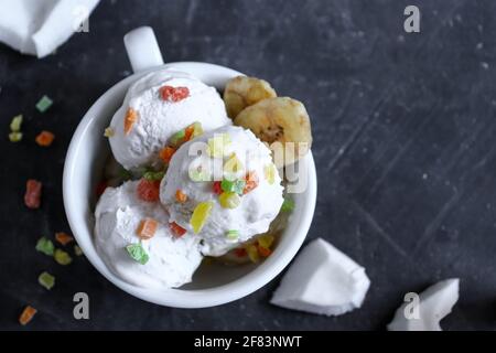 Coconut vegan ice cream with colorful fruit sprinkles. Sundae and candied fruits in a white cup. Dark background, copy space. Top view Stock Photo
