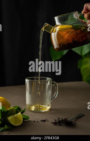 Woman pouring hot black tea with mint, lemon and lime from teapot into glass mug, evening tea party Stock Photo