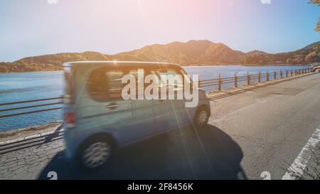 Eco car driving on road with lake and mountain landscape scenery. Motion blur showing car movement. Concept of road trip travel, rental cars, green Stock Photo