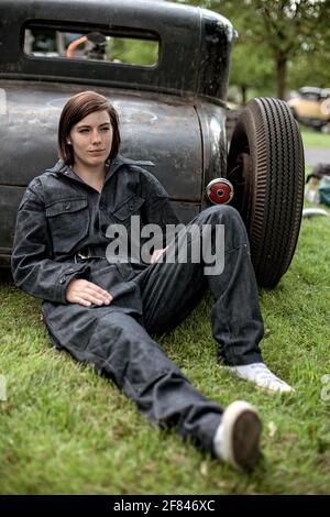 young beautiful woman in denim overall leaning on parked  hot rod car , Stock Photo