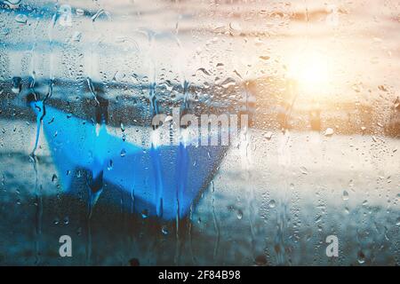 Blurry background with sunlight on water of blue paper boat washed up on beach after rain behind wet glass in raindrops Stock Photo