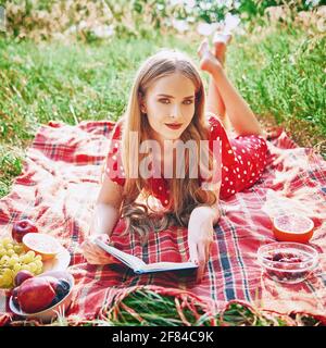 Picnic scene: attractive smiling girl lying on a plaid and reading book in grove. Outdoor portrait of beautiful young woman in red dress. Effect of vi Stock Photo