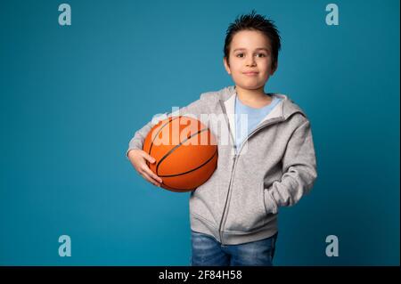 Portrait of an adorable sporty child holding a ball for playing basketball in hand, looking at camera and standing isolated over blue background with Stock Photo