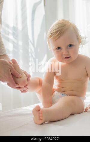 Mom gives her baby a leg and foot massage. Close-up. A satisfied baby is sitting on a massage table. Stock Photo