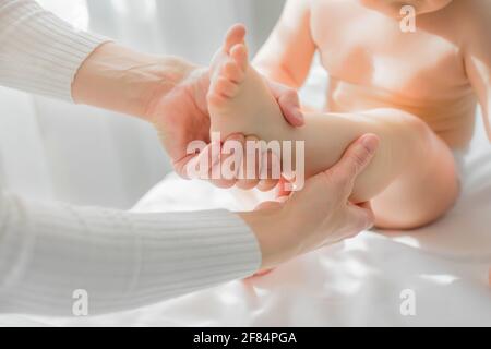 Mom gives her baby a leg and foot massage. Close-up. A satisfied baby is sitting on a massage table. Stock Photo