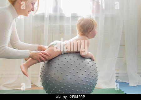 Mom is engaged in gymnastics with the baby on an inflatable ball. Fitball, gymnastics for a child. Stock Photo
