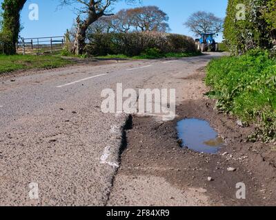 A blue farm tractor approaches surface damage to tarmac on a rural road is marked for repair with white spray paint. Taken on Wirral in the UK Stock Photo