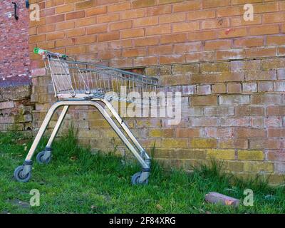 An unbranded, damaged supermarket shopping trolley discarded in an urban area. Pictured against a backdrop of a weathered brick wall. Stock Photo