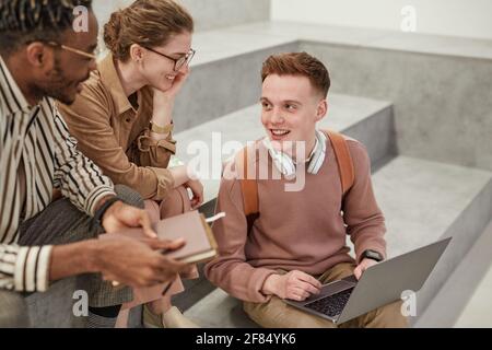 High angle view at group of cheerful students chatting indoors in modern college lounge, focus on young man using laptop with friends Stock Photo