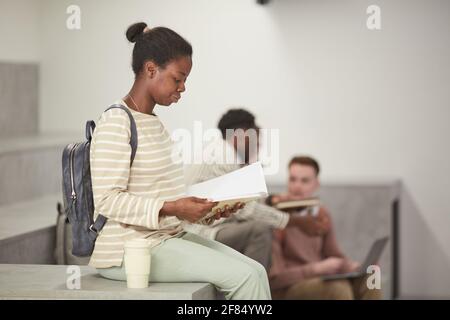 Side view portrait of young African-American woman reading books in modern college lounge, copy space Stock Photo