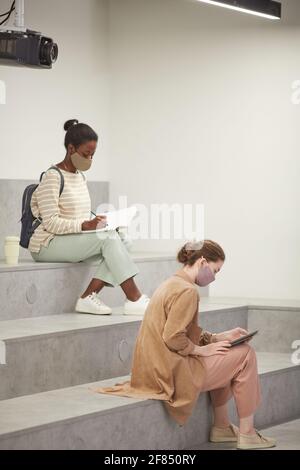 Vertical side view portrait of two young women wearing masks in school while studying in modern lounge Stock Photo
