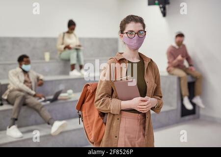 Waist up portrait of young female student wearing mask and looking at camera while standing in modern school lounge, copy space Stock Photo