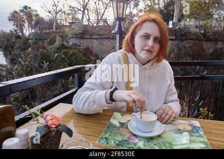 Terrace of restaurant by sea in Antalya, Turkey, young European red-haired woman relaxing at table with cup of coffee. Stock Photo