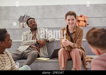 Diverse group of students chatting cheerfully while relaxing in modern school lounge, focus on smiling young woman talking to friend, copy space Stock Photo